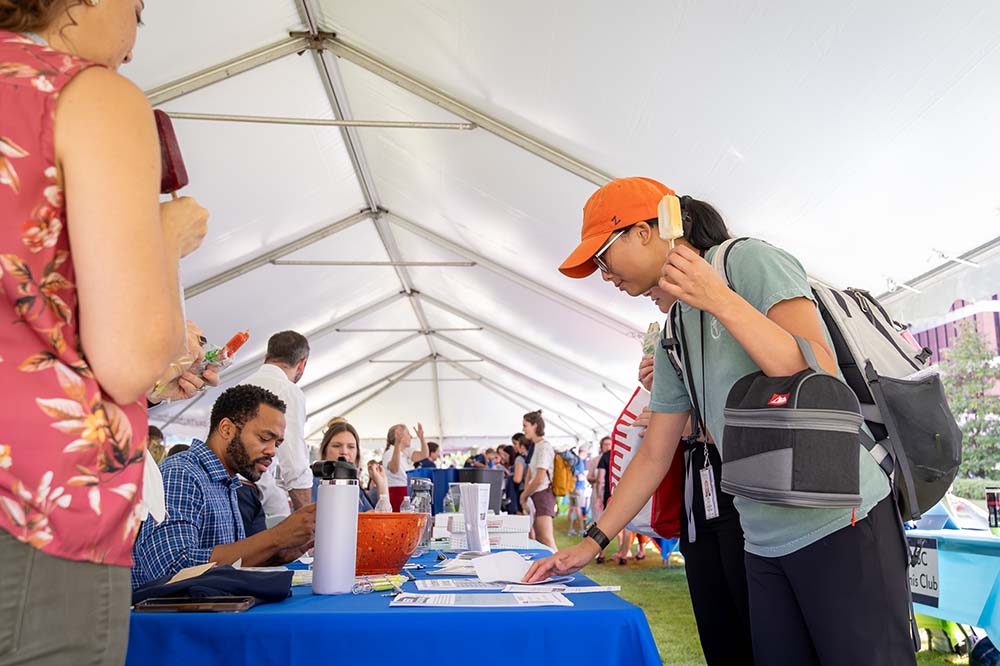 Person in an orange baseball cap leans over a table in a tent. A bearded man is seated at the table.